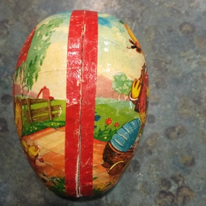May include: A vintage Easter egg made of papier-m&acirc;ch&eacute; with a red stripe down the centre. The egg is decorated with a colourful scene of a farm with a red barn, a fence, and a boy in a blue shirt and red hat.