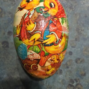 May include: A vintage Easter egg-shaped container featuring a colourful illustration of two chicks pushing a baby carriage with other chicks following behind. The chicks are yellow with orange beaks and black eyes. The background is green with a blue sky.