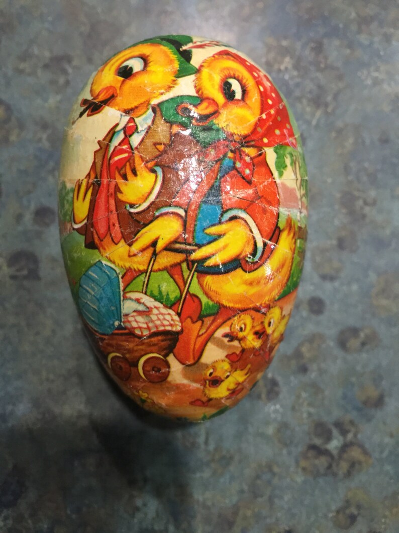 May include: A vintage Easter egg-shaped container featuring a colourful illustration of two chicks pushing a baby carriage with other chicks following behind. The chicks are yellow with orange beaks and black eyes. The background is green with a blue sky.