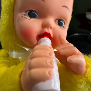 May include: A yellow plush toy baby doll with brown hair, blue eyes, and red lips is holding a white bottle to its mouth.