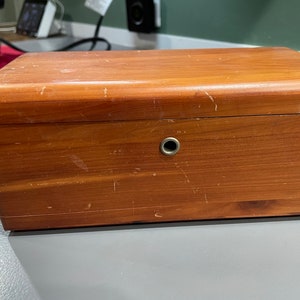 May include: A wooden box with a light brown finish and a small hole on the top. The box has a worn appearance with scratches and marks on the surface.