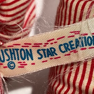 Puede incluir: Primer plano de una etiqueta blanca con costuras rojas y el texto "USHTON STAR CREATION" en letras azules con un s&iacute;mbolo de copyright.