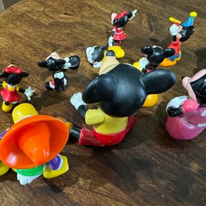 May include: A collection of nine plastic Disney character figurines, including Mickey Mouse, Minnie Mouse, and Goofy. The figurines are all in various poses and are painted in bright colors.