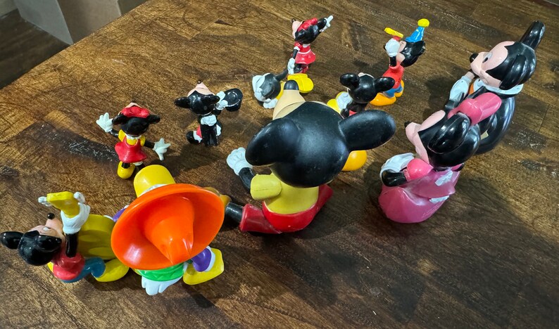 May include: A collection of nine plastic Disney character figurines, including Mickey Mouse, Minnie Mouse, and Goofy. The figurines are all in various poses and are painted in bright colors.