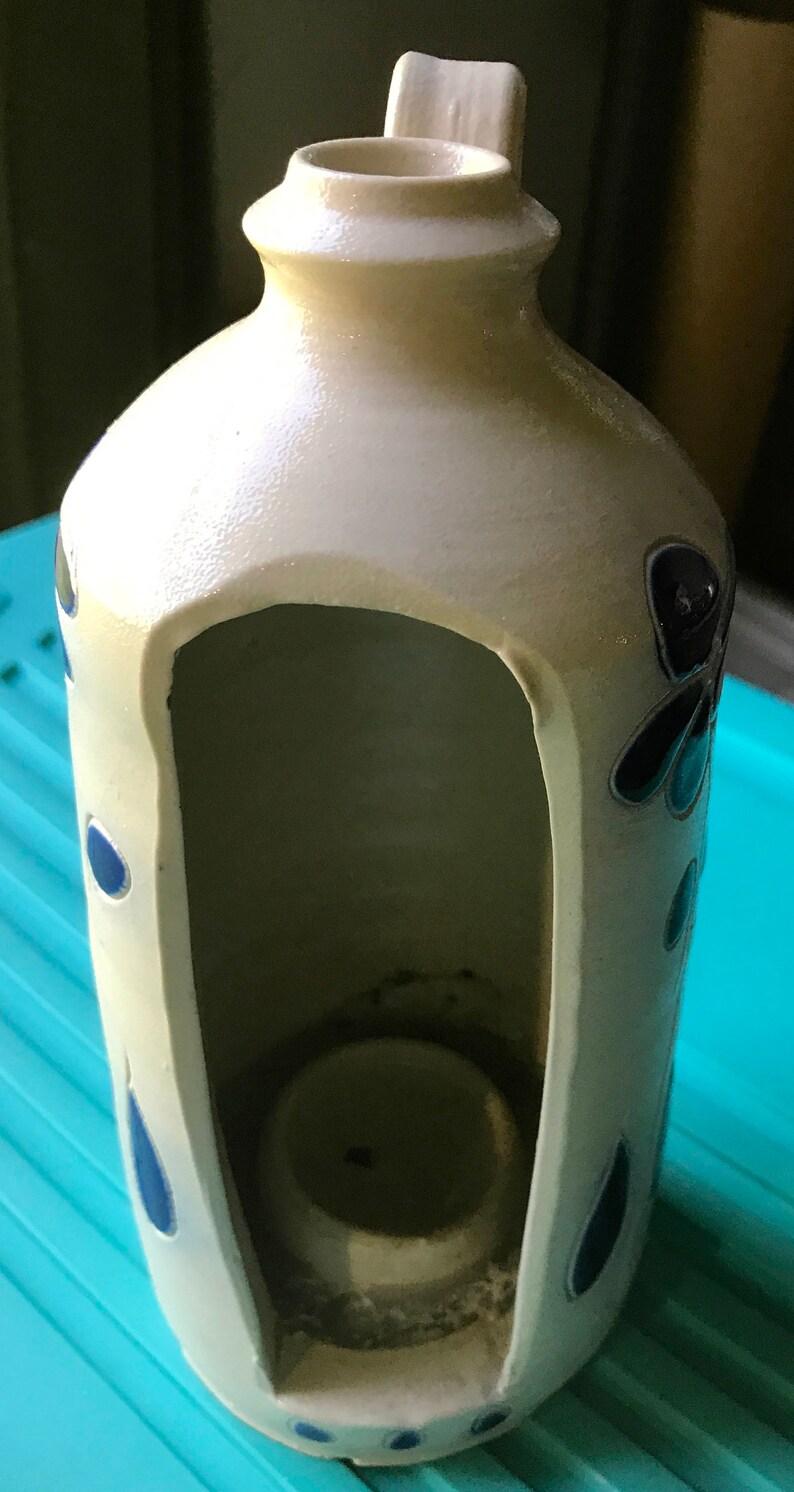 Puede incluir: Un portavelas de cer&aacute;mica blanca con un dise&ntilde;o floral azul y negro. El portavelas tiene un recorte en forma de casa con un peque&ntilde;o portavelas en el interior.