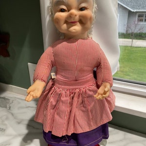 Puede incluir: Una mu&ntilde;eca vintage con una cara sonriente, cabello gris y un vestido a rayas rojas y blancas. La mu&ntilde;eca lleva pantalones morados y zapatos rojos.