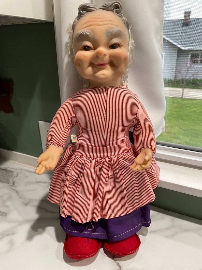 Puede incluir: Una mu&ntilde;eca vintage con una cara sonriente, cabello gris y un vestido a rayas rojas y blancas. La mu&ntilde;eca lleva pantalones morados y zapatos rojos.