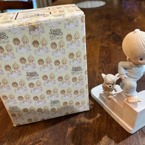 May include: A porcelain figurine of a boy and a dog climbing stairs. The boy is wearing blue pajamas and has blonde hair. The dog is white and brown. The figurine is on a white base with the words "Precious Moments" and "Love Forever" printed on it. The figurine is packaged in a box with a pattern of angels.