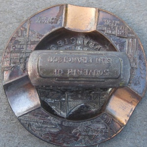 May include: A vintage brass ashtray with a built-in cigarette holder. The ashtray features a design of the Golden Gate Bridge and the text "Souvenir of San Francisco".