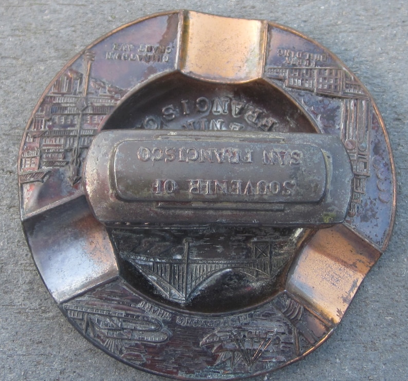May include: A vintage brass ashtray with a built-in cigarette holder. The ashtray features a design of the Golden Gate Bridge and the text "Souvenir of San Francisco".