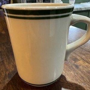 May include: A white ceramic mug with a green stripe around the top. The mug has a handle and is sitting on a wooden surface.