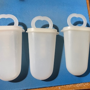May include: Three white plastic popsicle molds with a handle on top. The molds are on a blue surface.