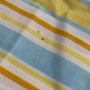 May include: A striped fabric with alternating bands of yellow, light blue, white, and orange. There are two small dark spots on the fabric.