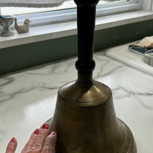 May include: A large, antique brass bell with a wooden handle. The bell has a worn, aged patina and a slight dent on the side.