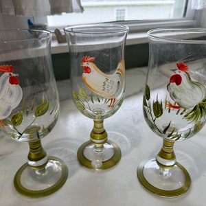 May include: Three clear glass wine glasses with gold and green painted stems and bases. Each glass features a hand-painted rooster design with green leaves.