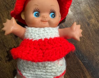 Könnte beinhalten: Eine Vintage-Puppe mit rotem gehäkeltem Hut, Kleid und Schuhen. Die Puppe hat ein weißes gehäkeltes Oberteil und eine Windel. Die Puppe hat blaue Augen und steht mit ausgestreckten Armen.