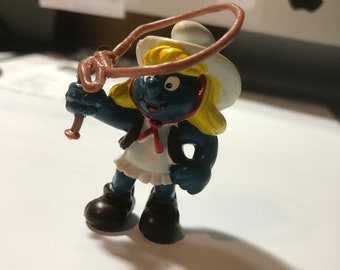 May include: A blue Smurf figurine wearing a white cowboy hat and a red bandana. The Smurf is holding a brown lasso in its right hand.