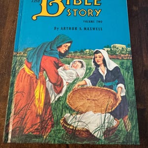 May include: The Bible Story, Volume Two, book cover. The cover is turquoise with the title in yellow and the author's name in white. An illustration depicts two women and a baby in a field with a basket. The women are wearing colorful robes.