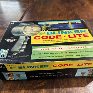 May include: Vintage Hasbro Twin Navy Blinker Code Lite set. The box features a graphic of a flashlight-style device, with text indicating it sends and receives Morse code. The box is black with yellow, blue, and green accents, and includes the text "Send Secret Messages".