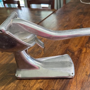May include: A vintage, silver-colored citrus juicer with a lever arm. The juicer has a bowl for catching juice and a base for stability. The lever arm is curved and extends over the bowl. The juicer is made of metal and appears to be well-used.