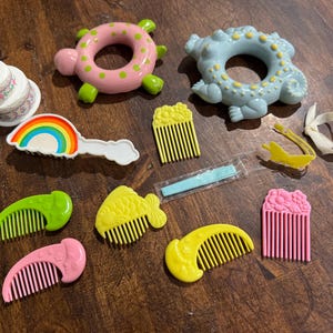 May include: A collection of colorful vintage hair accessories, including combs, a rainbow barrette, and a cake-shaped trinket. The accessories are in various shapes and colors, such as pink, green, yellow, and blue. The items are arranged on a dark wooden surface.