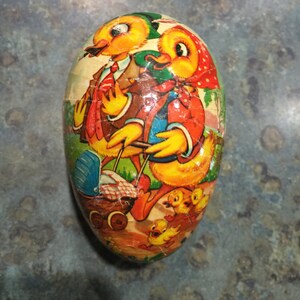 May include: A vintage Easter egg-shaped container featuring a colourful illustration of two ducks and their ducklings. The ducks are wearing clothes and the container has a yellow, orange, red, green, and blue colour scheme.