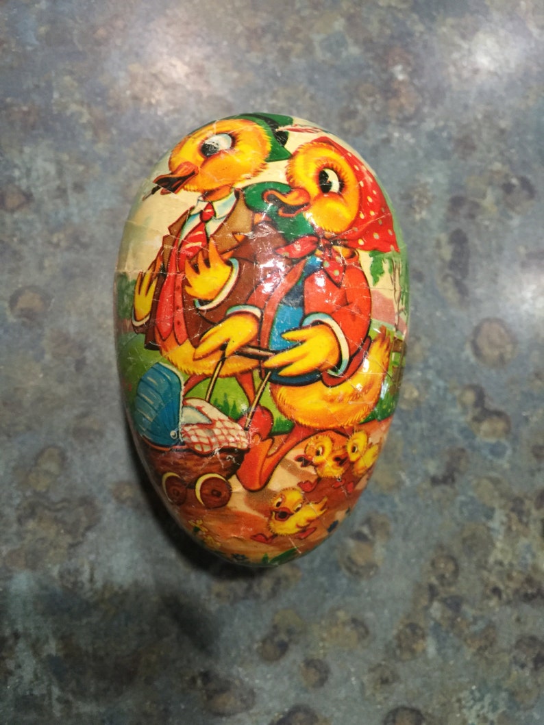 May include: A vintage Easter egg-shaped container featuring a colourful illustration of two ducks and their ducklings. The ducks are wearing clothes and the container has a yellow, orange, red, green, and blue colour scheme.