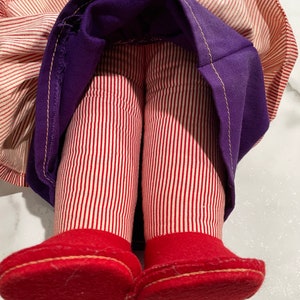 Puede incluir: Una mu&ntilde;eca vintage con piernas a rayas rojas y blancas y zapatos de fieltro rojos. La mu&ntilde;eca lleva una falda morada con una tela a rayas blancas y rojas debajo.