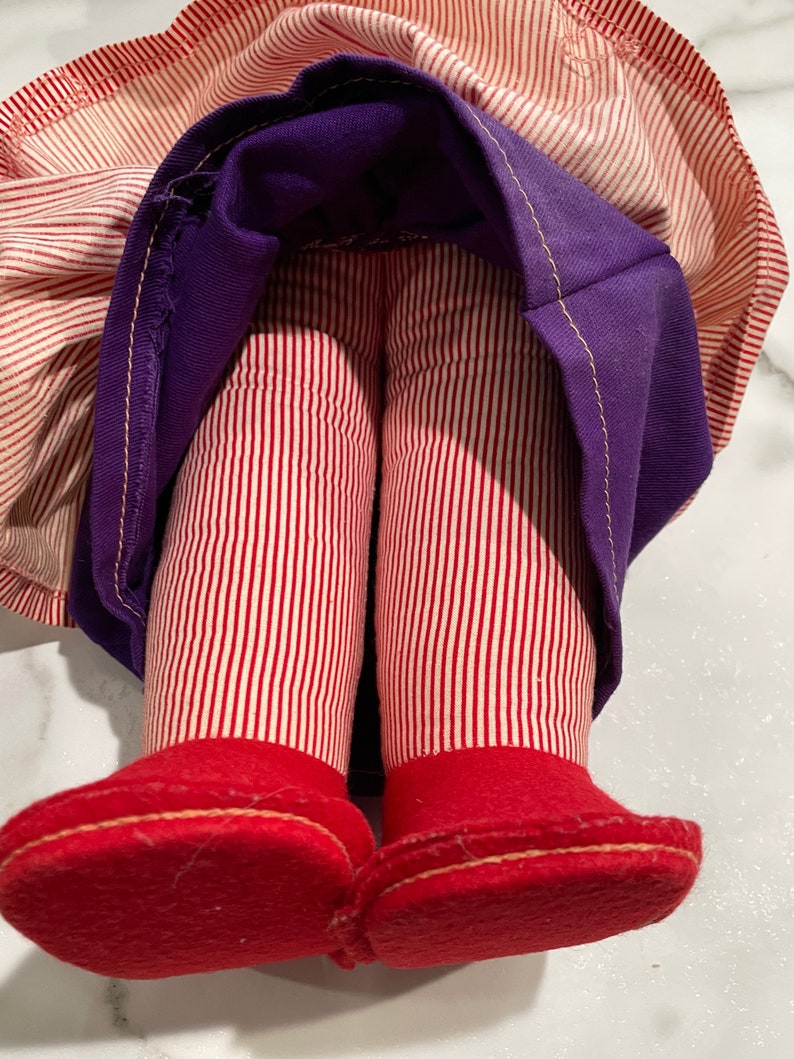 Puede incluir: Una mu&ntilde;eca vintage con piernas a rayas rojas y blancas y zapatos de fieltro rojos. La mu&ntilde;eca lleva una falda morada con una tela a rayas blancas y rojas debajo.