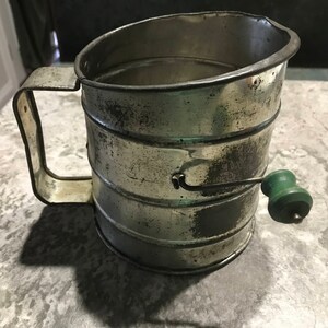 May include: A vintage silver metal flour sifter with a green plastic handle. The sifter has a handle and a crank to operate it.