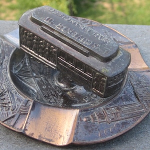 May include: A vintage metal ashtray featuring a San Francisco cable car with the text "SAN FRANCISCO CABLE CAR" and "SOUVENIR OF" on the top. The ashtray is decorated with a scene of the Golden Gate Bridge and other city landmarks.