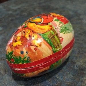 May include: A vintage Easter egg made of papier-m&acirc;ch&eacute; with a colourful illustration of chicks and a hen. The egg is decorated with a red ribbon and has a small opening at the top.