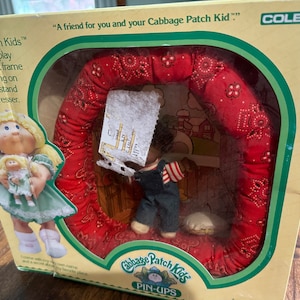 May include: A vintage Cabbage Patch Kids Pin-Ups doll set in its original packaging. The box features a doll with blonde hair and a green dress, and a small doll. The set includes a soft frame with a red bandana design.