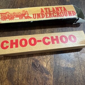 May include: Wooden toy train set with a box. The box is tan with red text that reads "ATLANTA UNDERGROUND" and a train graphic. The wooden block reads "CHOO-CHOO" in red letters.