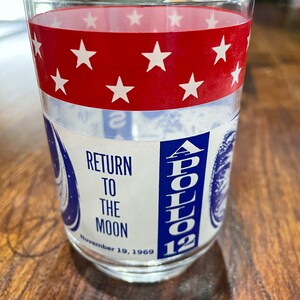 May include: A clear glass with a red and white band of stars and a blue and white label that says "Apollo 12 Return to the Moon November 19, 1969".