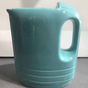 May include: A light blue ceramic pitcher with a wide mouth and a handle. The pitcher has a smooth, glossy finish and a slightly rounded shape.