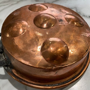 May include: Antique copper pan with a black iron handle. The pan has five round indents on the bottom.