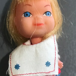 May include: A small plastic doll with blonde hair, blue eyes, and a white dress with blue stars. The doll is wearing a red and blue skirt underneath the dress.