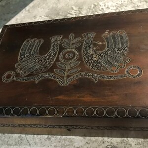 May include: A dark brown wooden box with a decorative design of two birds and a flower. The box has a raised edge with a series of circles around the perimeter.