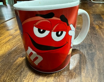 May include: A red ceramic mug with a cartoon red M&M's character with a mischievous expression. The character has a black mustache and eyebrows, and a white 'm' logo is visible on the mug.