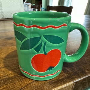 May include: Green ceramic mug with a red apple design and a red and white stripe around the top.