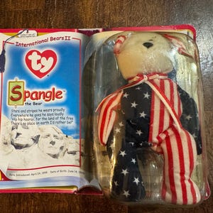 May include: A Ty Beanie Baby bear named Spangle, packaged in a clear plastic case. The bear is dressed in a patriotic outfit with a white head and a body featuring a blue background with white stars and red and white stripes. The packaging includes text.