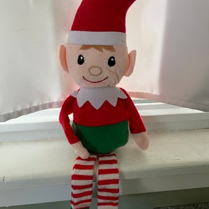 May include: A plush elf toy with a red hat, green pants, and white and red striped stockings. The elf is sitting on a windowsill.