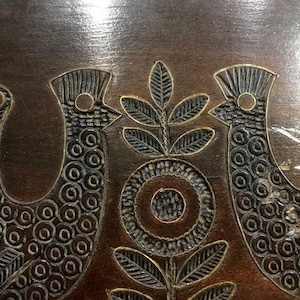 May include: A dark brown wooden surface with a carved design of two birds and a stylized flower. The birds are facing each other and have a circular pattern on their bodies. The flower has a circular center and four leaves.