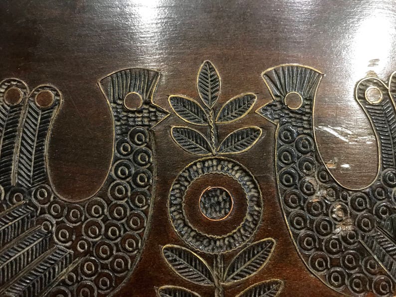 May include: A dark brown wooden surface with a carved design of two birds and a stylized flower. The birds are facing each other and have a circular pattern on their bodies. The flower has a circular center and four leaves.