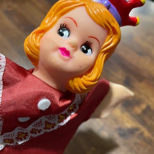 May include: A red and white doll with a crown, wearing a red dress with white lace trim and white polka dots. The doll has blonde hair and a painted face with large eyes.