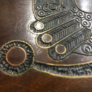 May include: A close-up of a wooden box with intricate metal inlay. The inlay features a circular design with a copper center and a black and gold border. The box is decorated with a stylized bird design.