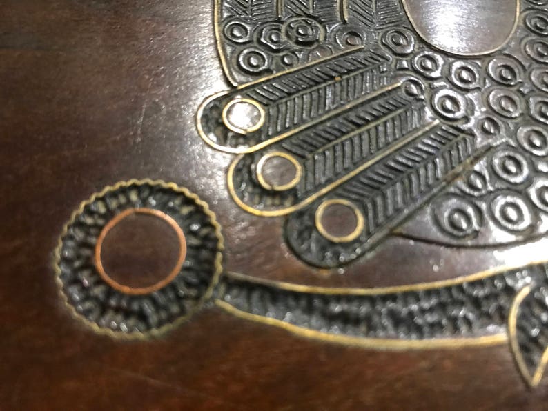 May include: A close-up of a wooden box with intricate metal inlay. The inlay features a circular design with a copper center and a black and gold border. The box is decorated with a stylized bird design.
