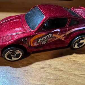 May include: A metallic red toy car with black wheels and a rear spoiler. The car has the text "zero G" in orange and white on the side. A fun collectible toy car.