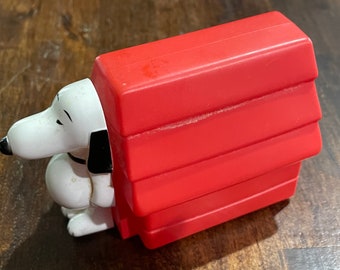 May include: A white plastic toy dog with black ears and a black collar, peeking out from a red plastic doghouse.